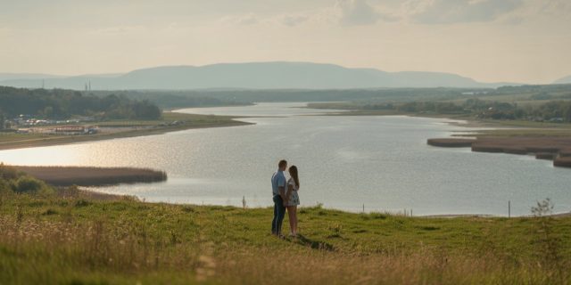 Para na wzgórzu o zachodzie słońca patrzy na rozległe jezioro i dalekie pasma gór, spokojny krajobraz.